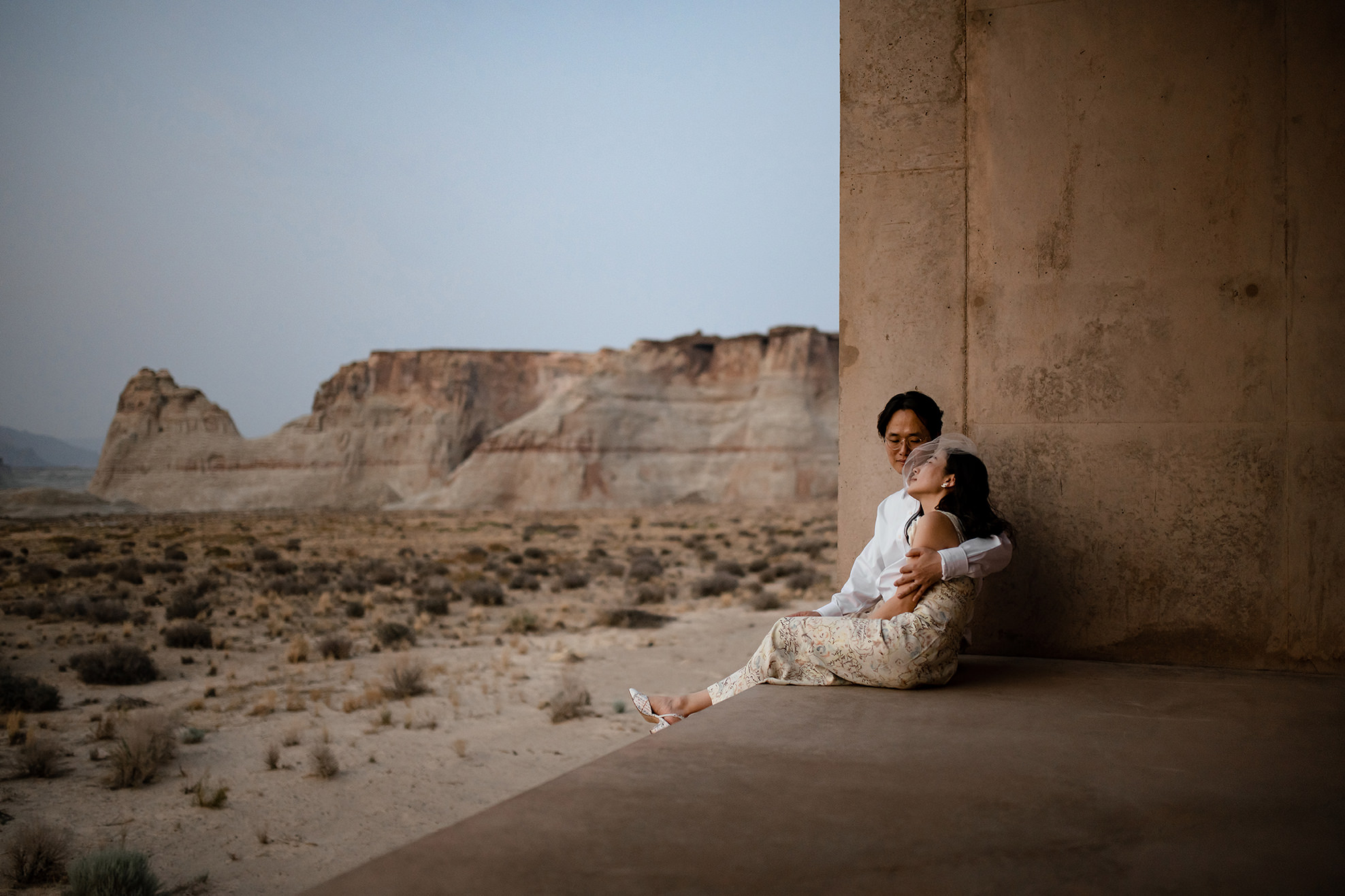 Husband holds his wife around the shoulder while they sit on a concrete structure in the dessert.