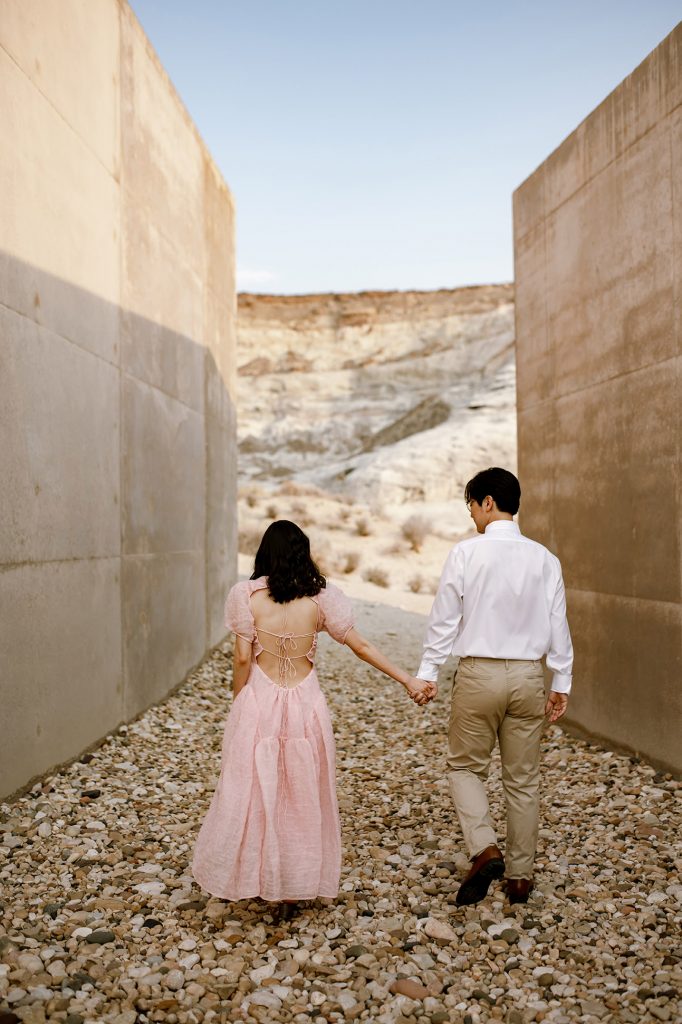 Couple holding hands walking on a rock path between two huge cement walls.