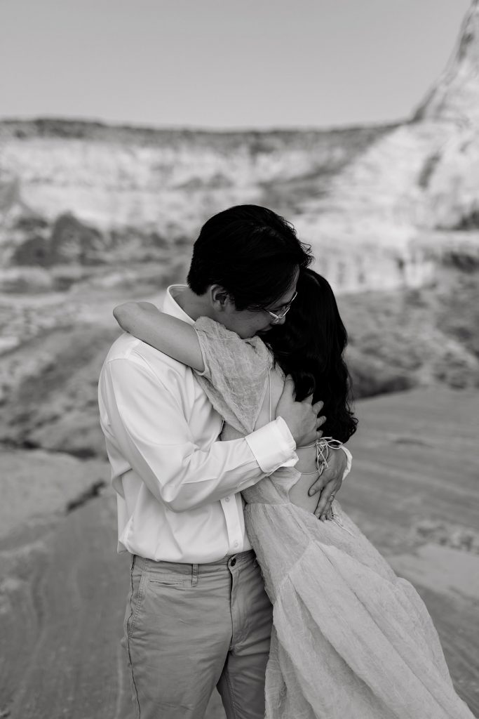 Black and white of husband and wife hugging each other with massive dessert bluffs surrounding them.