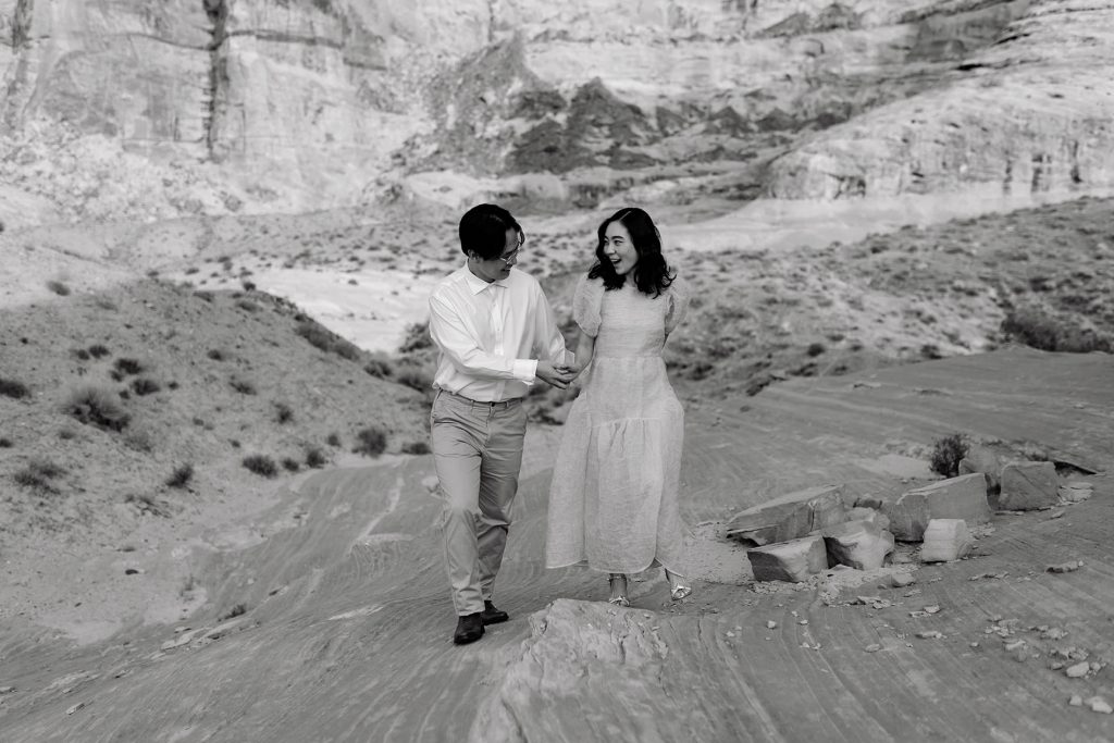 Husband and wife hold hands and laugh as they walk up a rocky desert hill in black and white.