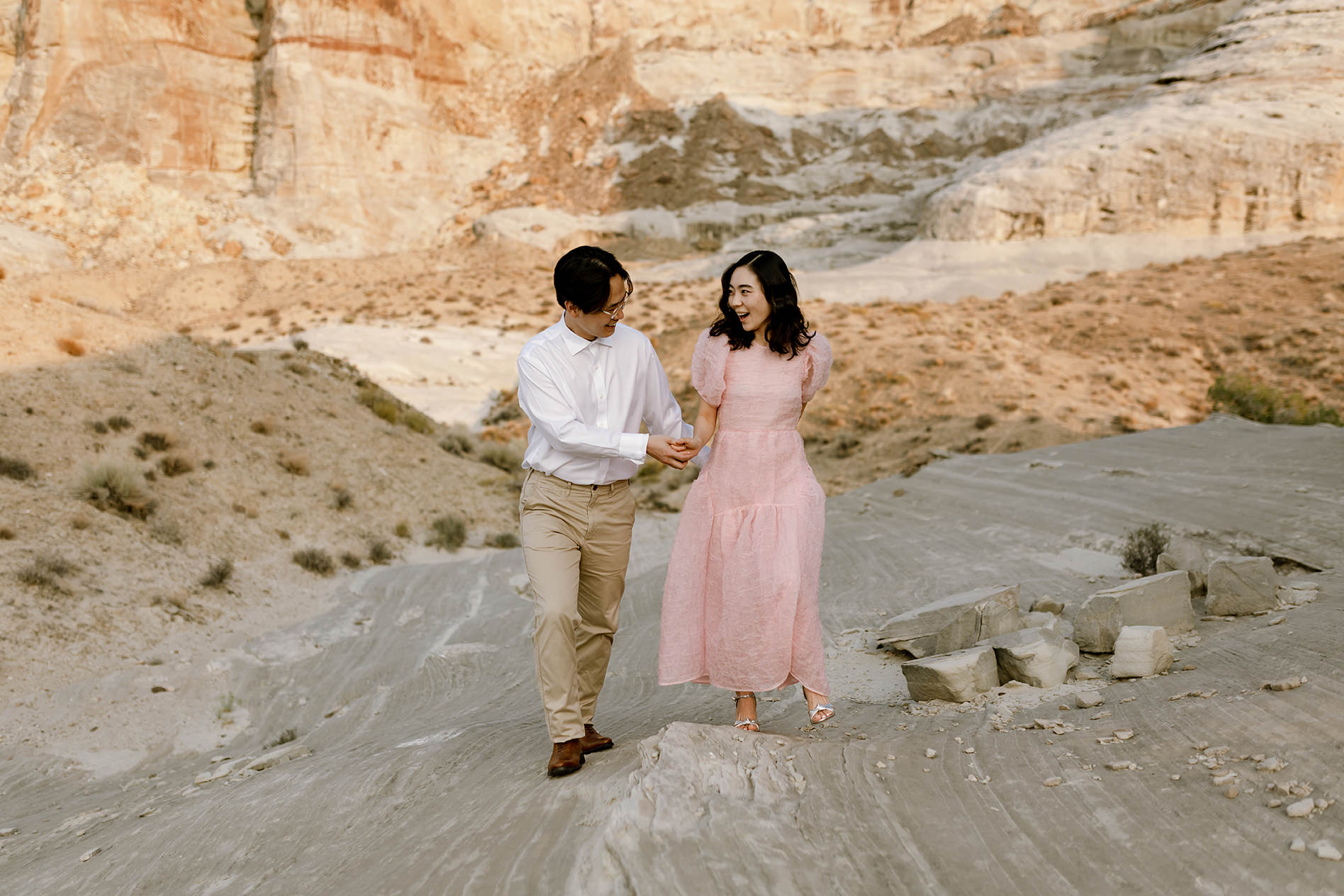 Husband and wife hold hands and laugh as they walk up a rocky desert hill.