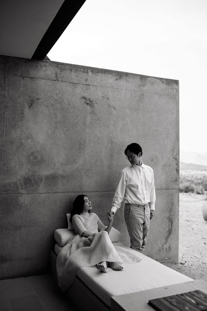 Husband stands and holds his new wifes hand while she lays under a blanket on their patio.