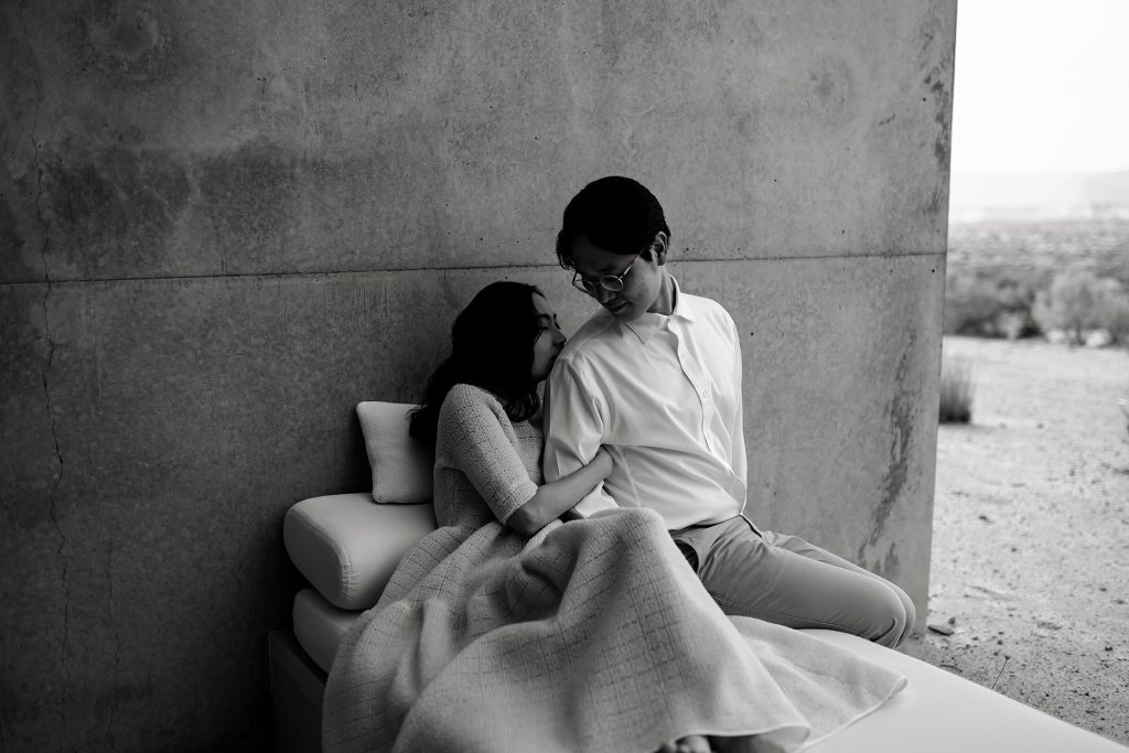 Husband and wife embrace eath other, cuddled up with a blanket on their patio.