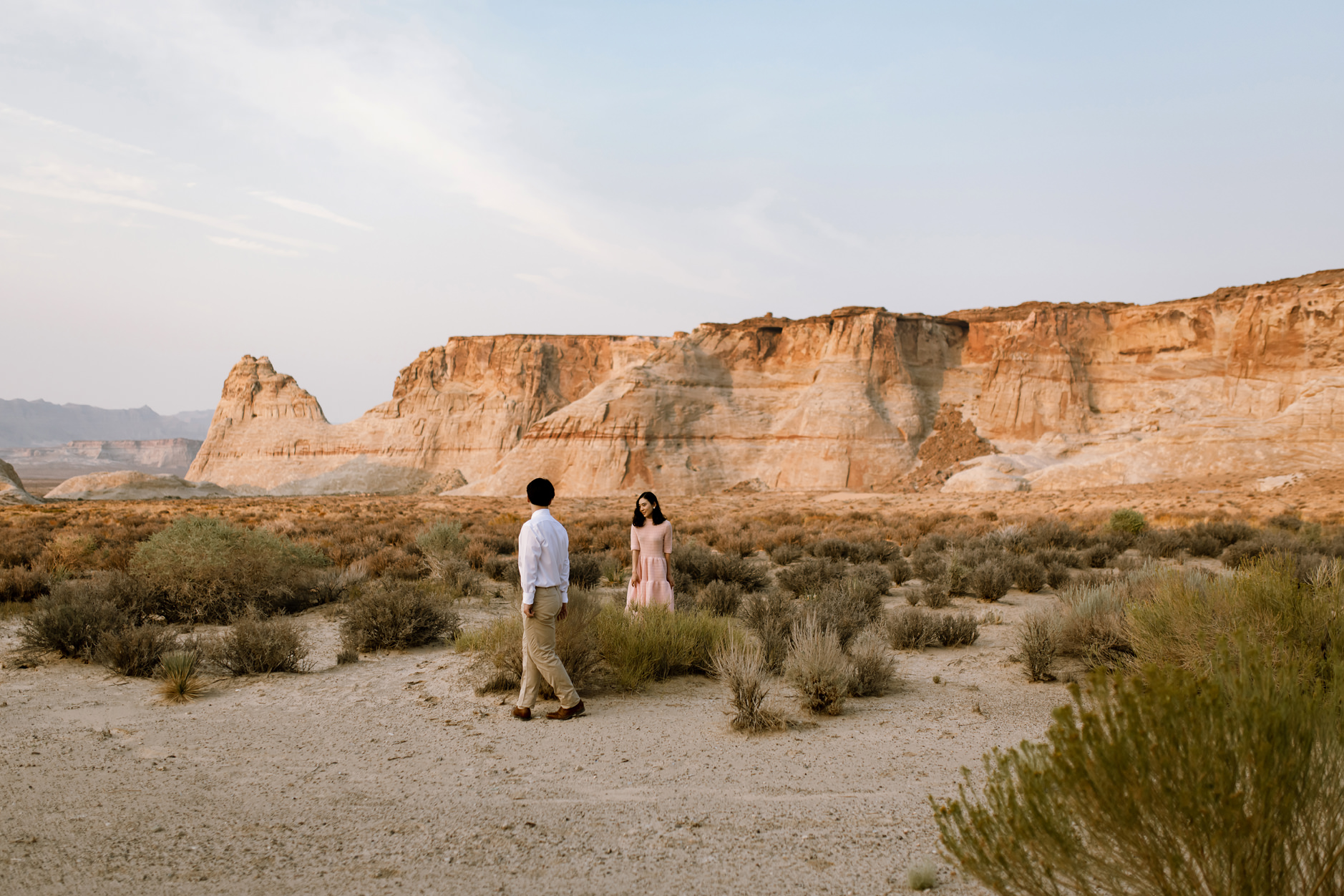 Husband walks around waist high desert brush towards his wife wearing a pink dress.