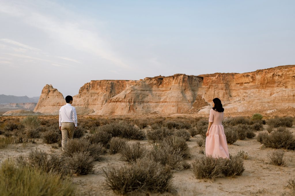 Man and woman walk out towards a dessert plateau, through the brush, while staying a small distance from each other.